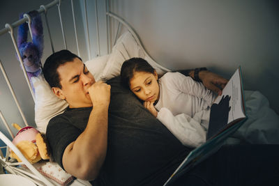 High angle view of father reading book to daughter while lying in bed at home