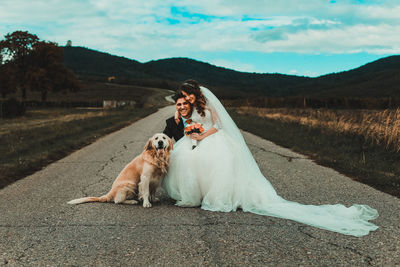 Bride and groom sitting by dog on road