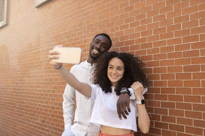 Young man and woman taking selfie through smart phone in front of brick wall