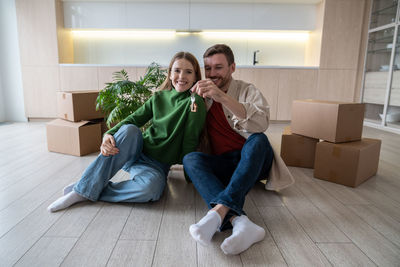 Family couple holding keys from new rented apartment sitting on floor among cardboard boxes.