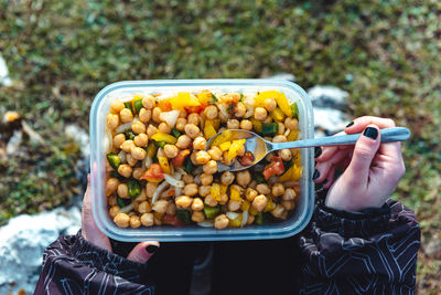 From above crop woman in warm jacket holding plastic container of chickpea with veggies having lunch on meadow while hiking in spain