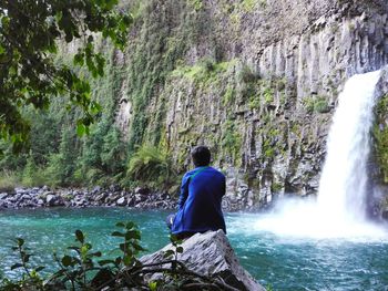 Rear view of man sitting on rock by river against waterfall in forest