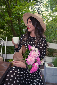 Smiling young woman with coffee and bouquet sitting at porch 