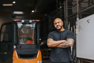Portrait of smiling bald mature male worker standing with arms crossed at warehouse