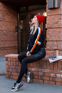 Full length of woman sitting on bench against brick wall outdoors