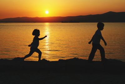 Silhouette siblings walking at beach against sky during sunset