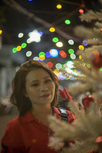 Portrait of young woman standing against illuminated christmas tree at night