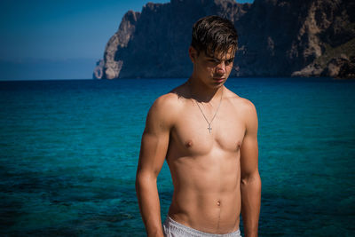 Shirtless young man standing at beach against sky
