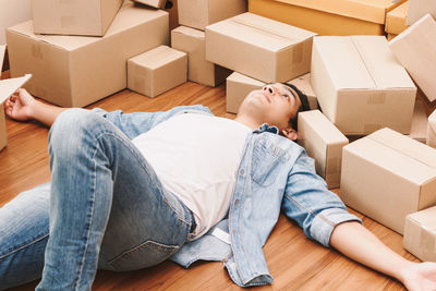 High angle view of young man lying by cardboard boxes on floor at new home