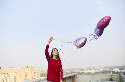Smiling woman with heart shape balloon standing against clear sky