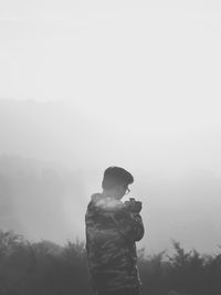 Rear view of man photographing on field against sky in foggy weather