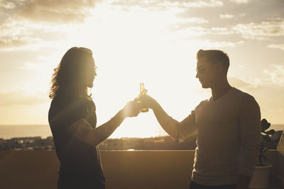 Male friends toasting drinks while standing against sky during sunset