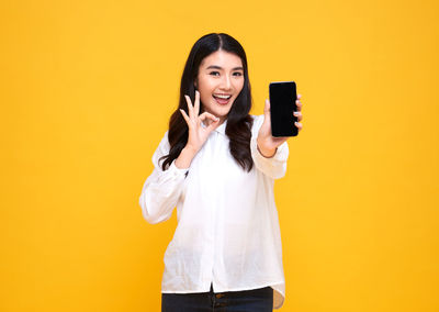 Young woman using phone while standing against yellow background