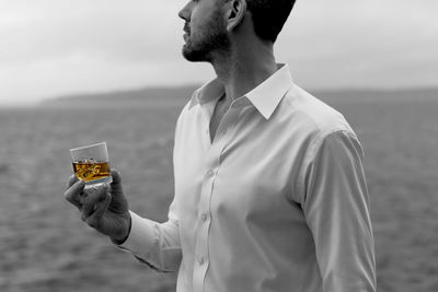 Midsection of man holding whisky in glass while standing on shore