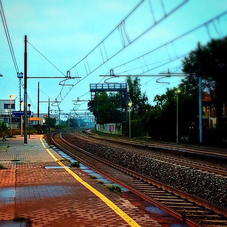 railroad track, rail transportation, transportation, public transportation, railroad station platform, railroad station, railway track, clear sky, built structure, building exterior, sky, tree, architecture, train - vehicle, power line, the way forward, train, electricity pylon, diminishing perspective, city