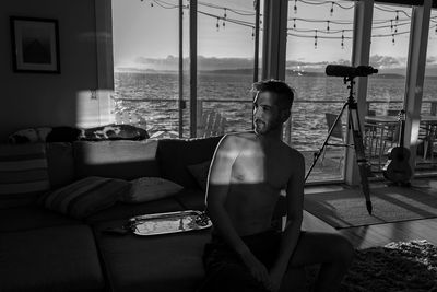 Sunlight falling on shirtless man looking away while crouching at home against sea