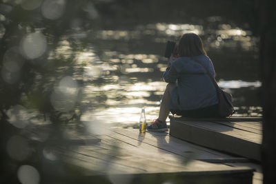 Rear view full length of woman photographing while sitting on pier against lake