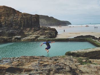 Man jumping on rock by sea against sky