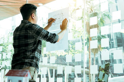 Side view of young man standing against window