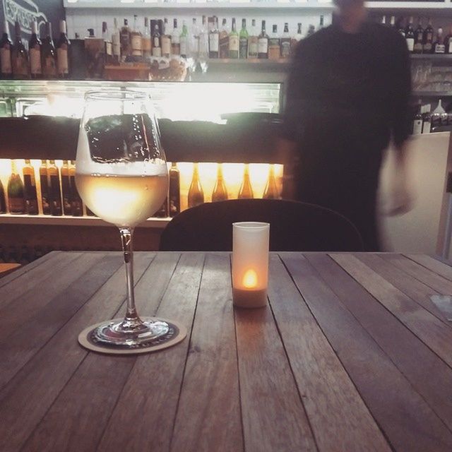 table, indoors, illuminated, restaurant, wood - material, candle, drink, food and drink, focus on foreground, night, drinking glass, lighting equipment, incidental people, alcohol, close-up, refreshment, glowing, lit, wooden, chair