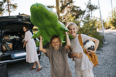 Portrait of happy siblings holding soccer ball and crocodile swim float while standing on driveway