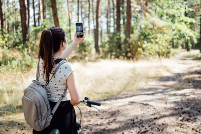 Young woman with backpack riding bike and taking photo on cell phone on pine forest background