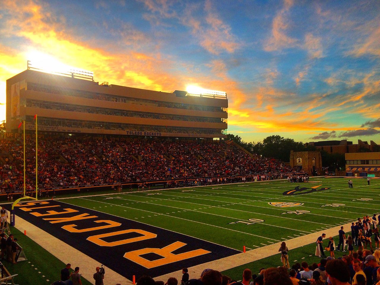 sunset, large group of people, architecture, city, building exterior, high angle view, built structure, sunbeam, stadium, cloud - sky, city life, sky, yellow, crowd, outdoors, office building, vibrant color, dramatic sky