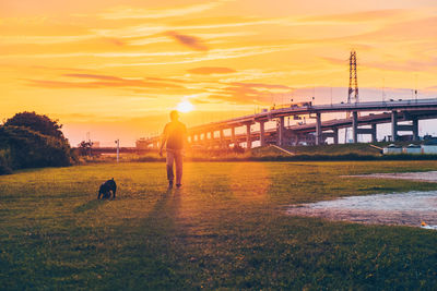 Man walking with dog on grassy field against orange sky