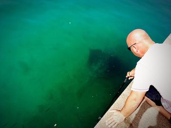 High angle view of mature man fishing in sea while kneeling on retaining wall