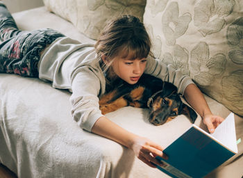 Girl reading book while lying on sofa with dog