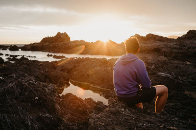 Rear view of man standing on rock against sky during sunset