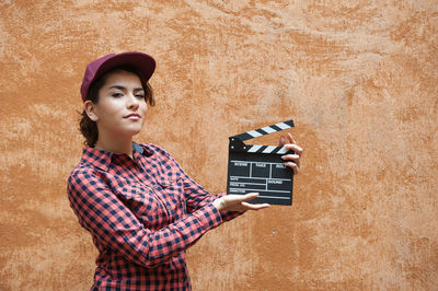 Portrait of young woman holding film slate against wall