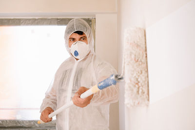 Full length portrait of man working with paint roller in protective suit and mask against wall