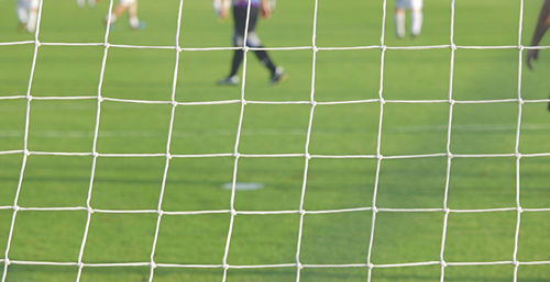 Low section of men playing soccer on field seen through goal post