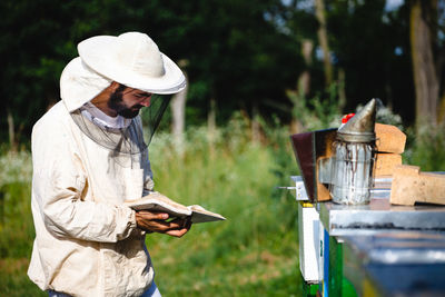 Side view of beekeeper with book examining beehive on land