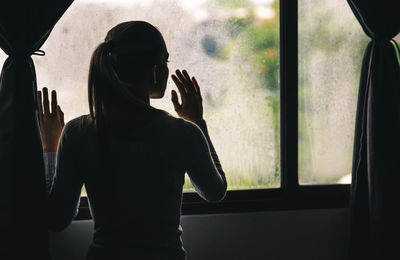 Rear view of silhouette woman standing by window at home