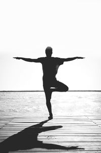 Full length of silhouette man standing on one leg on pier over sea against clear sky