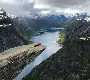 Scenic view of woman peeking over edge of trolltunga in norway at lake, snow, mountains, waterfalls