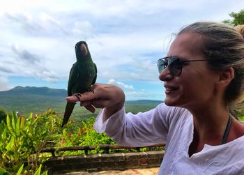 Smiling woman with parrot on finger against sky