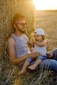 Father and son in t-shirts sitting next to a haystack on a sloping field during sunset