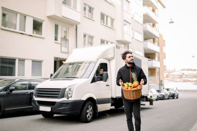 Young delivery man carrying fruit basket on street in city