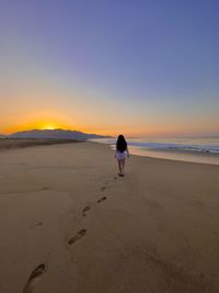 Rear view of woman walking at beach against clear sky during sunset
