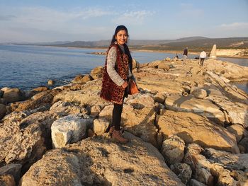 Full length portrait of woman standing on rock at beach against sky