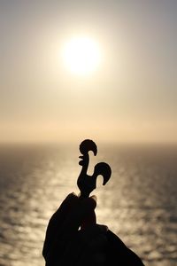 Cropped image of person holding silhouette rooster figurine by sea against sky during sunset