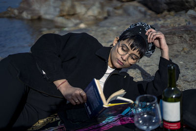 Young beautiful indian girl reading a book while leaning down on the ground wearing a black coat.