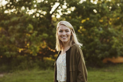 Portrait of smiling young blond woman standing against trees