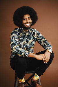 Portrait of smiling man sitting on stool against brown background