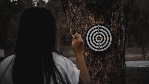 Rear view of woman holding dart against target on tree trunk in forest