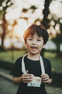 Portrait of happy boy with toy camera standing at park during sunset