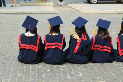 Rear view of students wearing graduation gowns while sitting on footpath